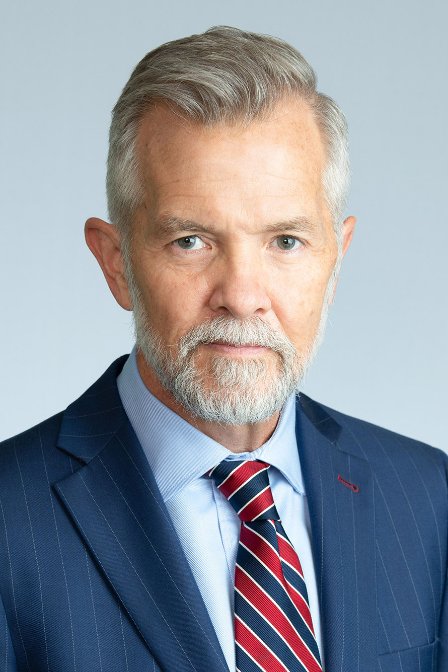 Photo: David Chard, a white man with short gray hair and short gray beard. He wears a blue suit and maroon tie. He poses in front of a light blue background.