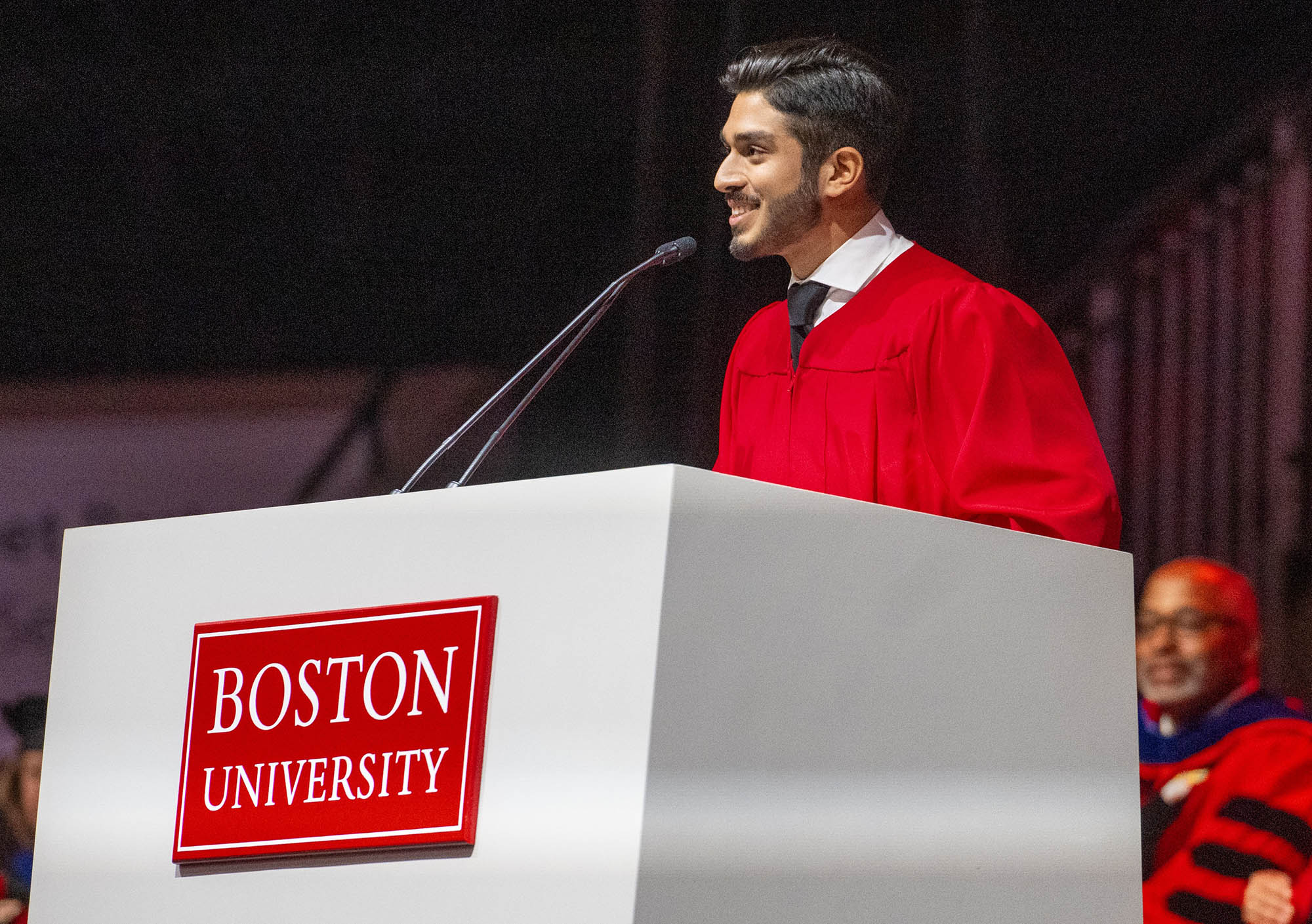 Photo: Dhruv Kapadia, a young tan adult man wearing red ceremonial robes speaks at a large white podium with a red sign that reads "Boston University".
