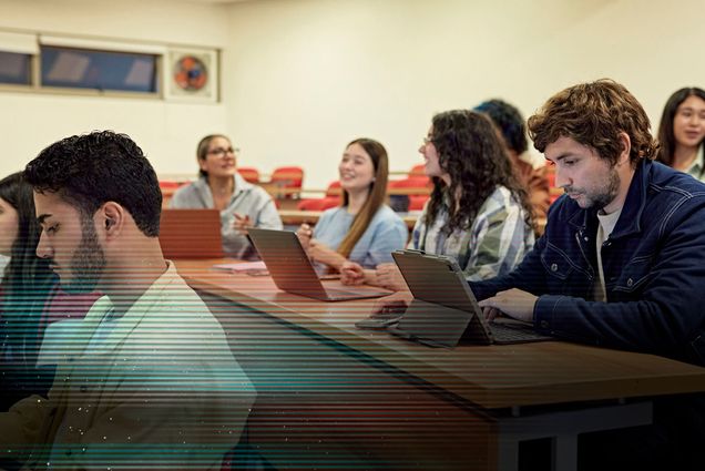 Photo: College students sit in a tiered college classroom as they work on laptops and chat with each other.