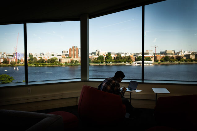 Photo: A shadowed student looks down at a table as they study in the renovated study lounge at Kilachand Hall. Behind them,. large windows feature a view of the Boston city skyline and the Charles River.