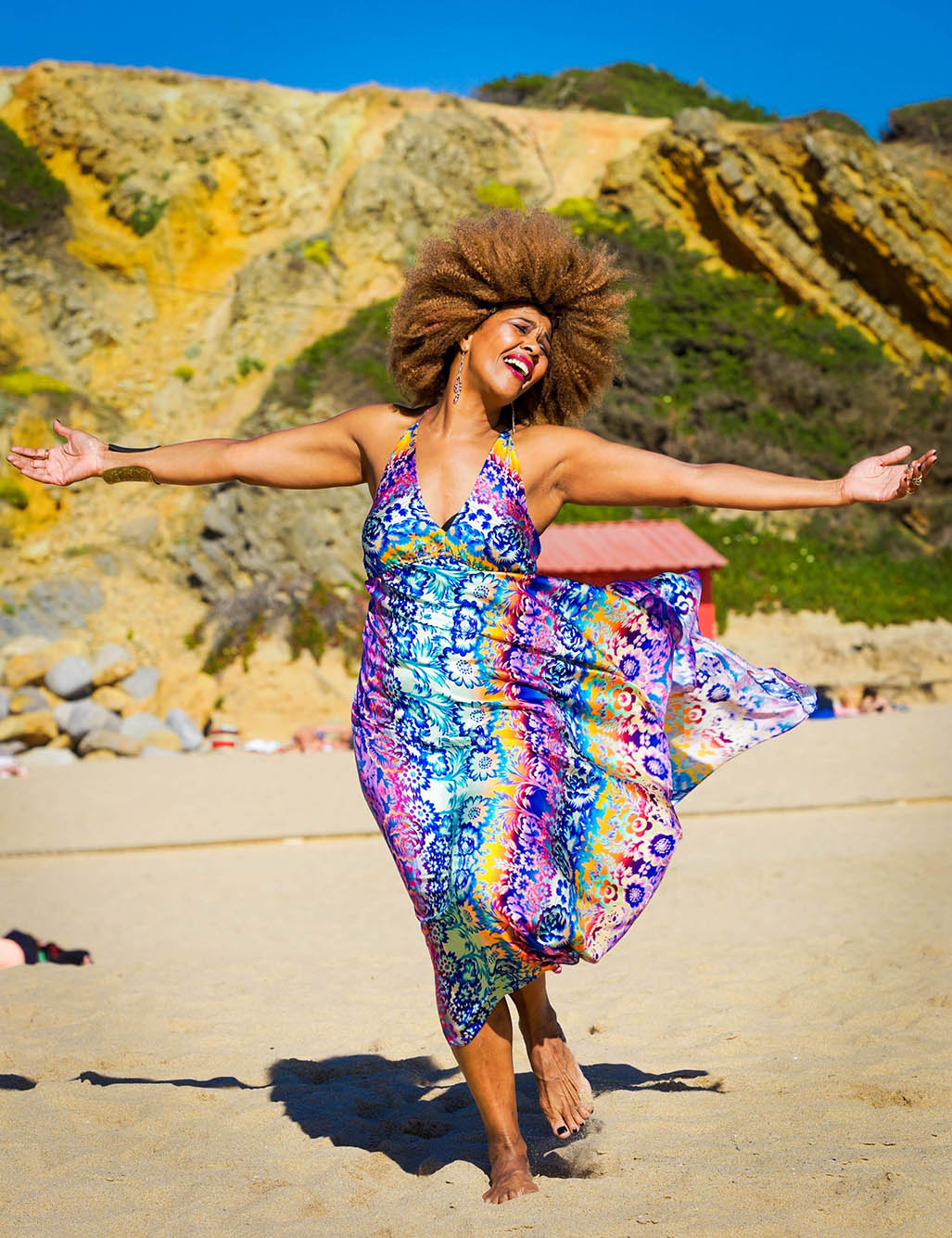 Photo: a Cape Verdean woman with tan skin and large, dark blonde afro poses with arms outstretched. She wears a rainbow dresses that flows behind her as she poses on a scenic beach.