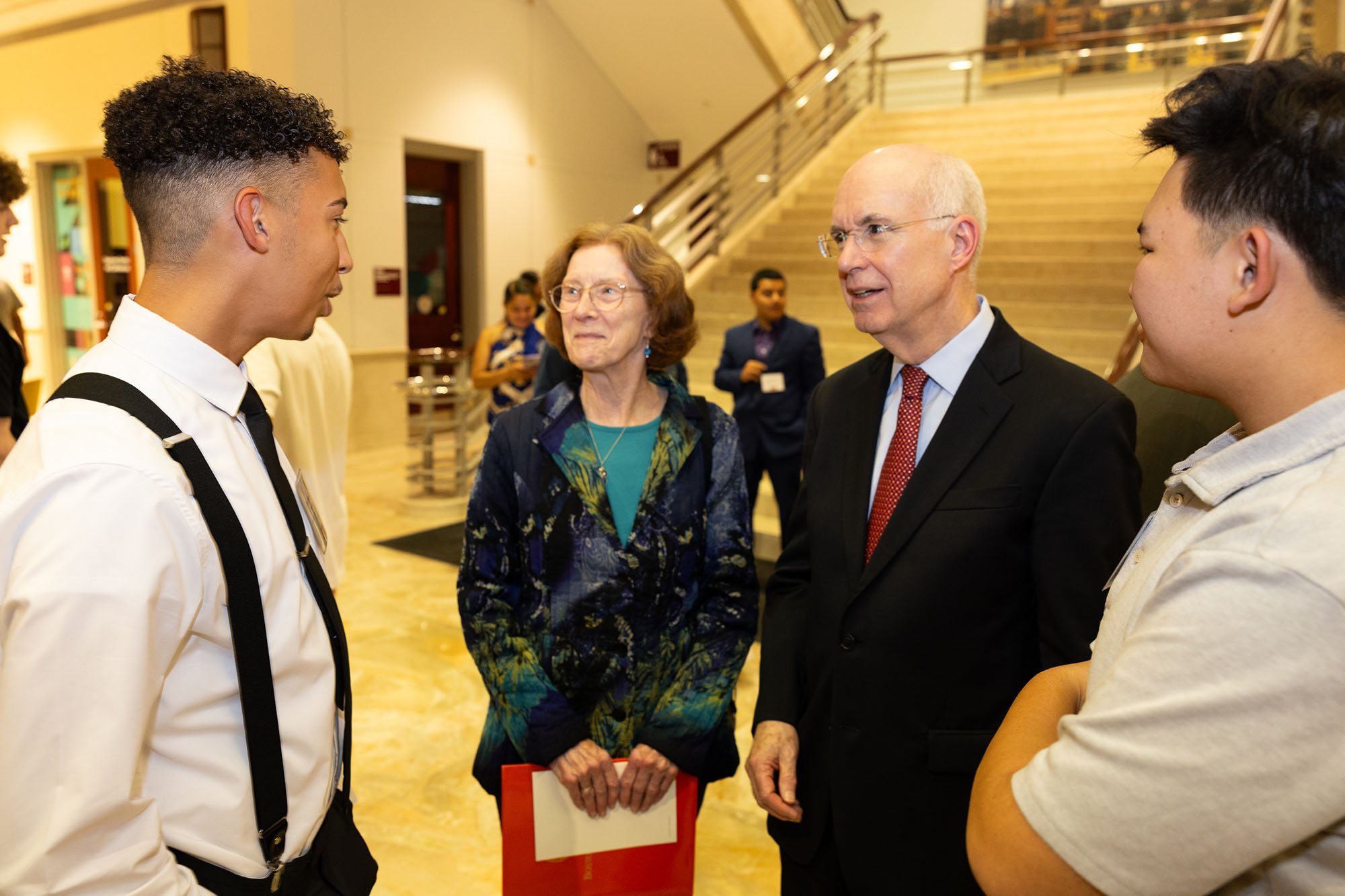 Photo: At last Thursday’s welcome reception, Kenneth Freeman (right), an older white man with white hair and wearing glasses, a light blue, sleeved collared shirt, red tie, and black jacket and pants, chats with two smiling students wearing business casual wear (left).