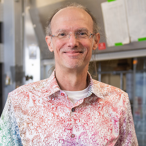 Photo of Mark Grinstaff, a middle-aged white man with glasses who wears a button-down shirt with a gradient, squiggly line pattern that fades from green and blue to red and pink. He smiles as he stands and leans against a cluttered lab table.