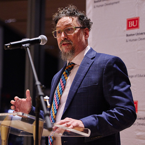 Photo: Action shot of Hank Fien, a man with short brown hair and bear wearing glasses, a white collared shirt, red and blue striped tie, and blue blazer. He stands at a glass podium in front of a white "Boston University" labeled backdrop.