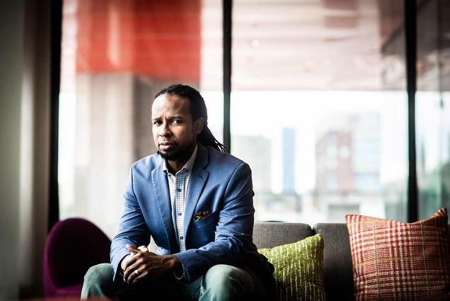 Photo of Ibram X. Kendi, a Black man with with long dreadlocks sits in a blue suit looking out a window.