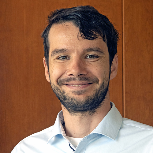 Photo: Headshot of Ignaty Leshchiner, a man with short black hair and wearing a light blue collared shirt, smiles and poses in front of a wooden background.