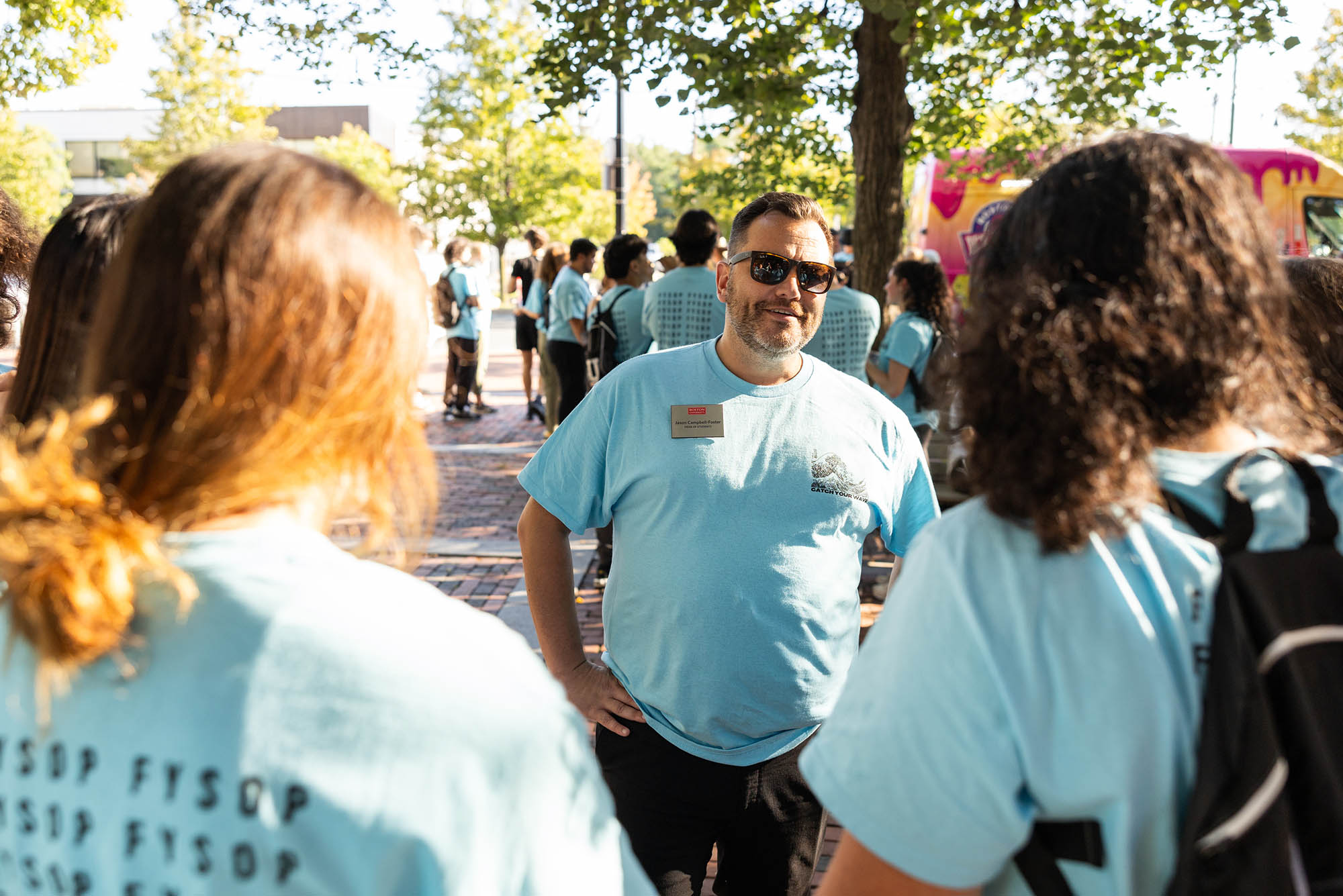 Photo: Jason Campbell-Foster, a white man with gray hair and a graying beard, stands with students outside with a blue t-shirt. He is smiling and standing with his hands on his hips.