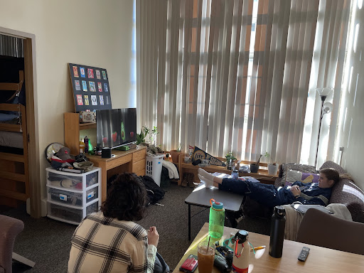 This is a photo of a Boston University campus living space. There are white shades that are blocking the sunlight from entering through the large windows in the corner of the apartment. There is a couch, a desk, two tables, two TV's, and plenty of water bottles and clothing scattered across the room. The carpet is grey and towards the left of the photo is a doorway to what appears to be someones bedroom. There is something playing on the TV. There are two individuals in the. One is on the couch and has on an all navy outfit with grown hair. They have their feet up on the coffee table. The person to the left is in a chair and has on a white and black flannel. Their brown hair is mostly covered by a beanie and their back is turned towards the camera.