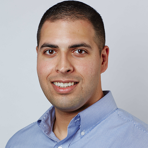 Photo: Headshot of Lou Awad. A man with a shaved head and wearing a light blue collared shirt smiles and poses for a photo in front of a light background.