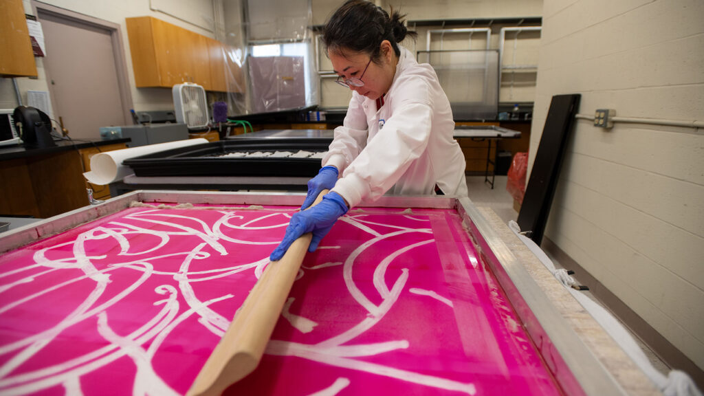 Photo: Lucy Kim, an ?Asian woman wearing glasses, a white lab coat, and purple gloves, works on spreading substance on a large pink screen print with large white squiggles on it. She works in a small lab area.