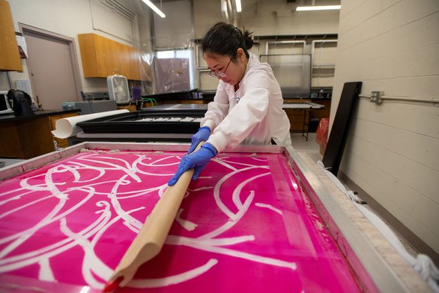 Photo: Lucy Kim, an ?Asian woman wearing glasses, a white lab coat, and purple gloves, works on spreading substance on a large pink screen print with large white squiggles on it. She works in a small lab area.