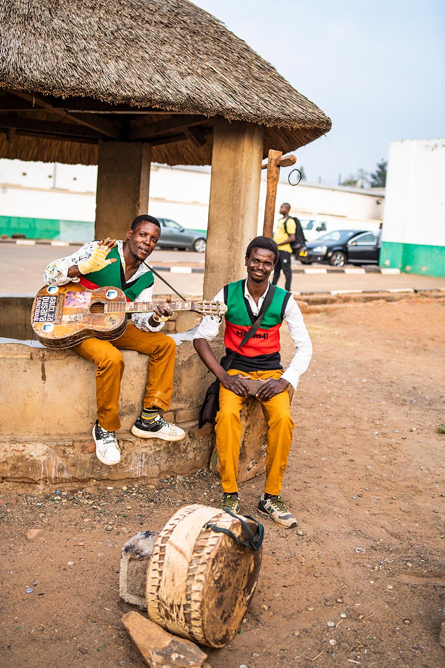 Photo: Two people wearing matching outfits pose for a photo at a Malawian roadside. They wear white collared shirts, red, green, and black sweater vests, and khaki pants.