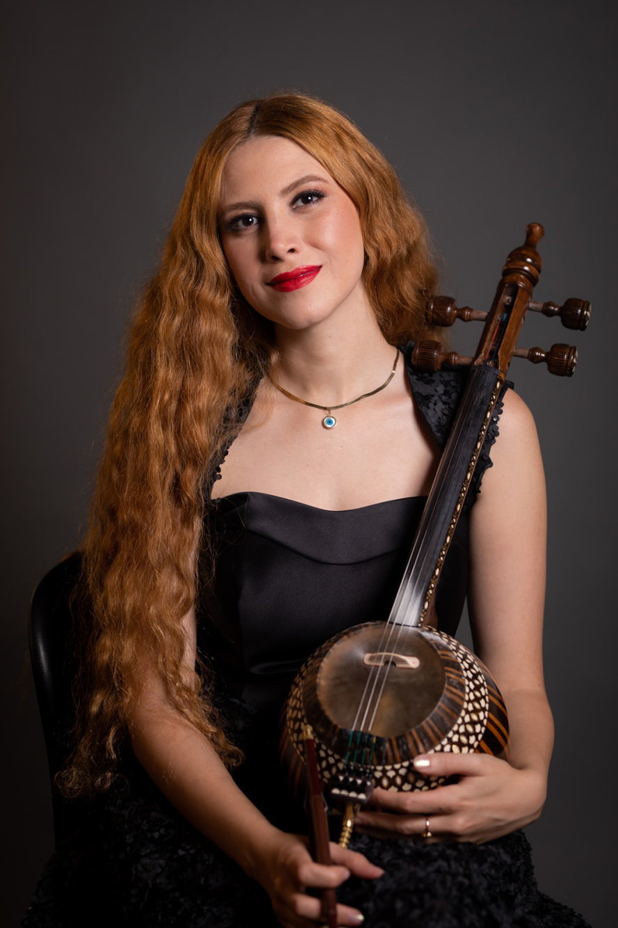 Photo: A white woman with long red hair and wearing a black dress poses with a kamancheh (a stringed instrument) in her arms. She poses in front of a black background.