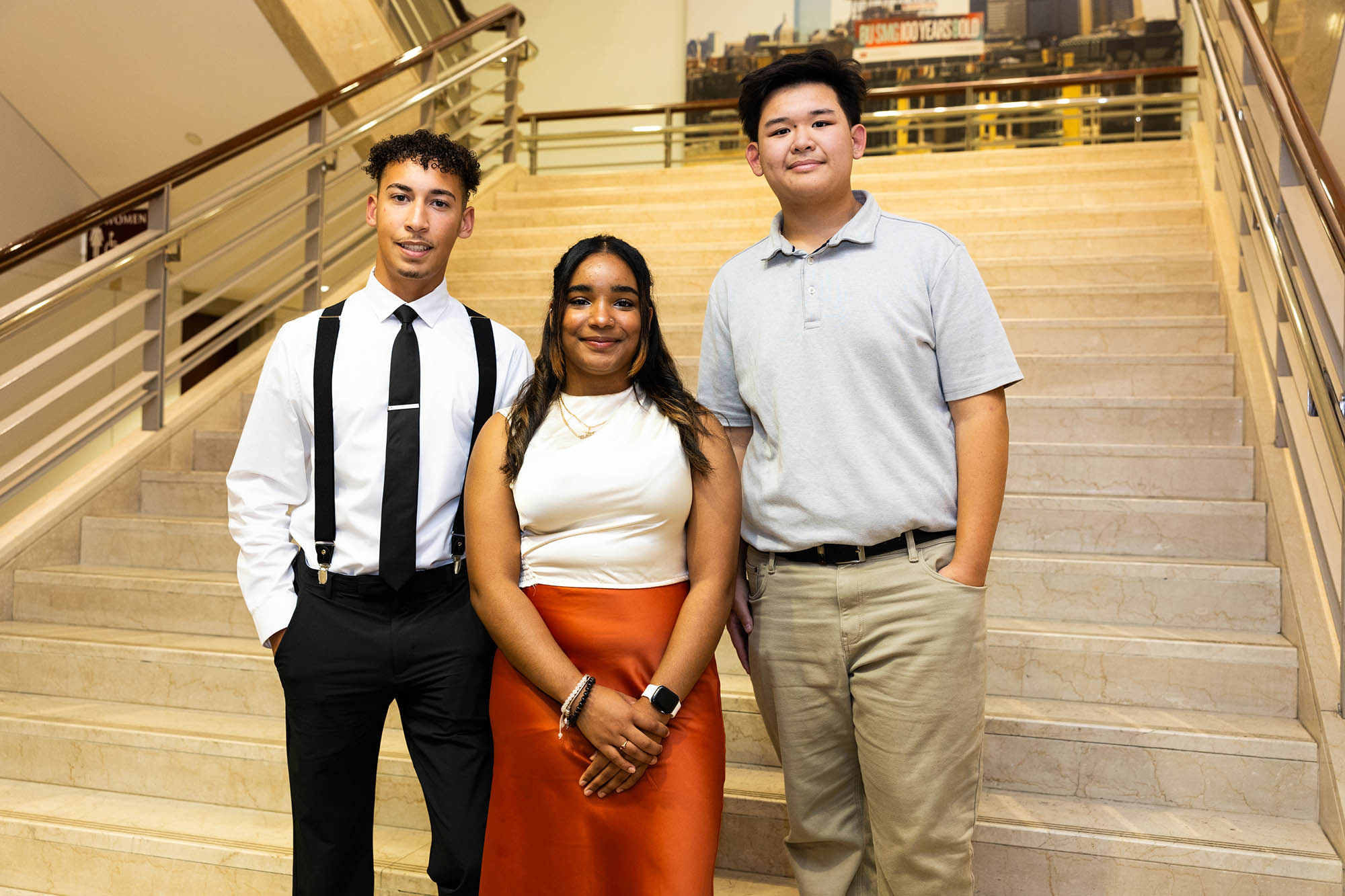 Photo: Three students wearing business casual attire stand and pose at the bottom of a large set of stairs. From left to right: Zaki Araujo, a tan young man with black, curly hair and wearing a light blue collared shirt and black tie, suspenders, and pants. Raysa Mendoza, a young, tan woman wearing a white blouse and long, red skirt. Kenny Phan, a young man wearing a light blue collared shirt and khaki pants.