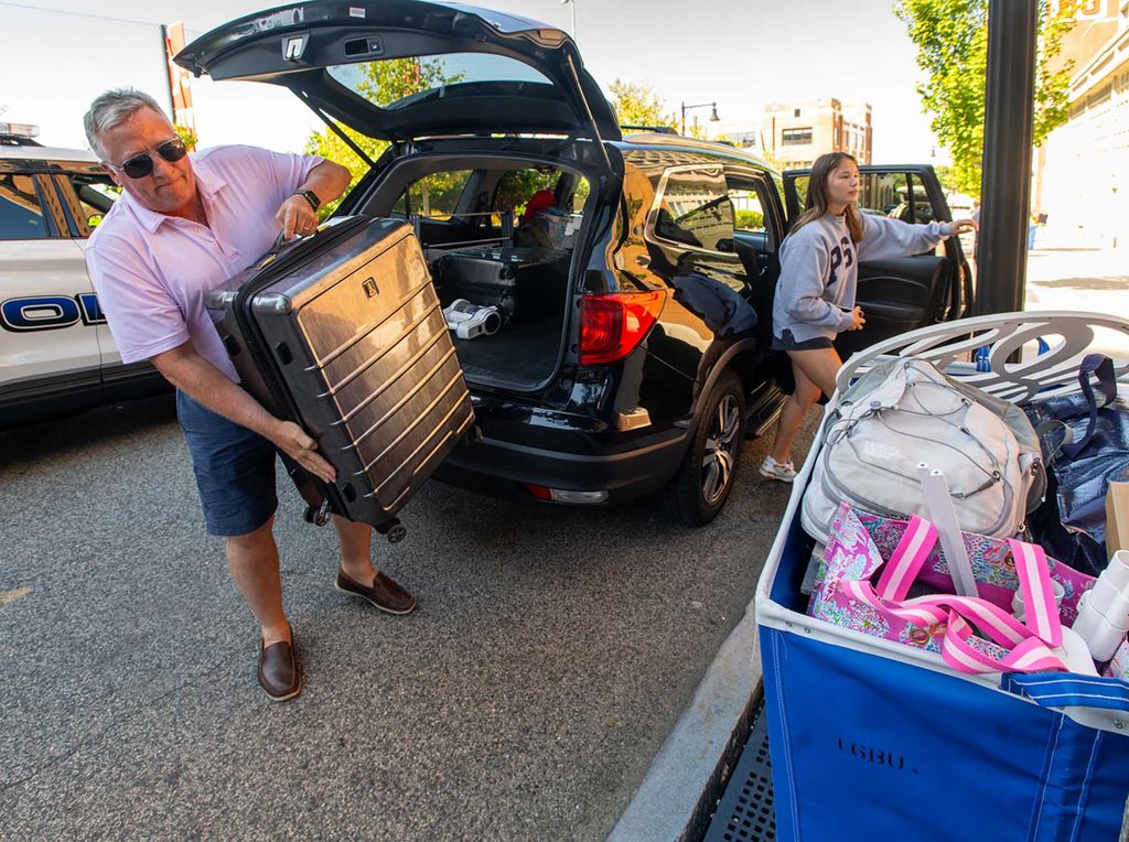 Photo: An older man wearing a purple shirt and jean shorts pulls a large suitcase out of the back of a car.