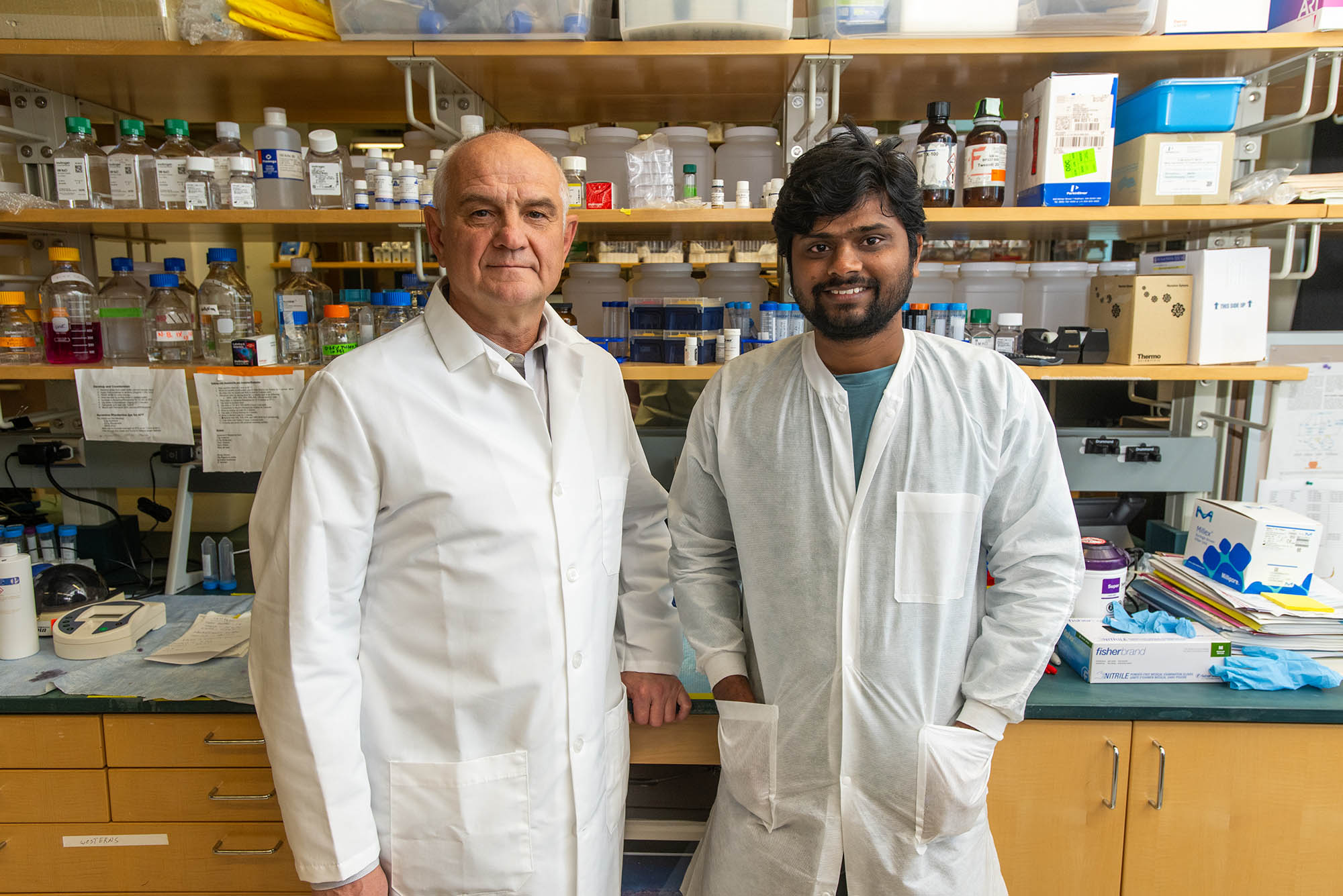 Photo: Igor Kramnik (left), an older white man wearing a white lab coat, and Shivraj Yabaji (right) a tan man with black hair and beard wearing a white lab coat, smile and pose in a science lab.