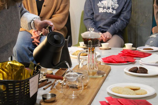 Photo: A table full of various tea accessories is shown. A person to the left is shown carefully pouring hot water into a glass teapot filled with tea leaves, as others around the tables prepare other teas and cups.