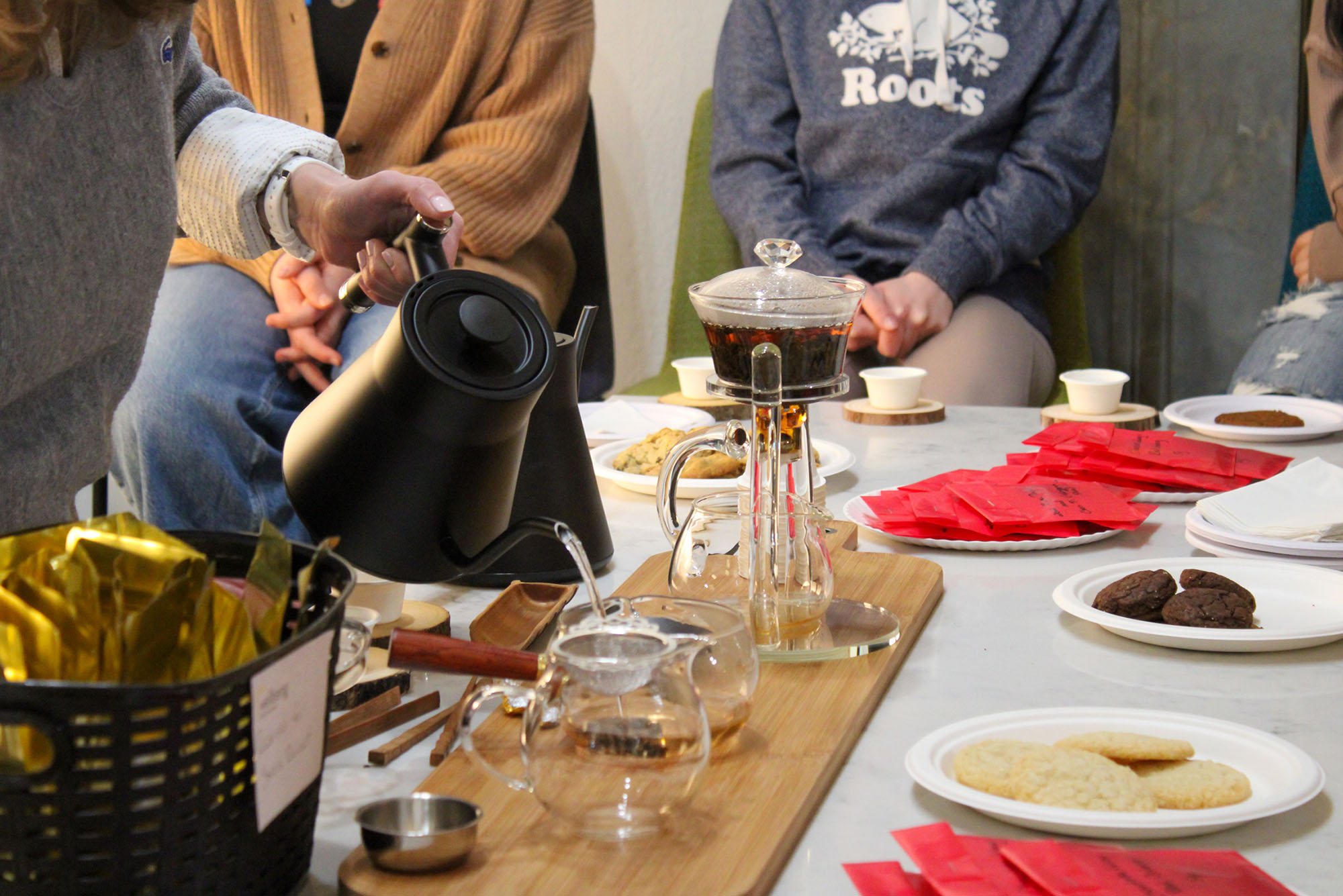 Photo: A table full of various tea accessories is shown. A person to the left is shown carefully pouring hot water into a glass teapot filled with tea leaves, as others around the tables prepare other teas and cups.