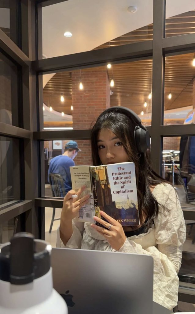 This is a photo of Robin Kim (COM'24) reading a book in a coffee shop. She has brown hair and headphones on and the book is covering half of her face. In front of her, there is an open laptop and a water bottle. The book she is reading is titled The Protestant Ethic and the Spirit of Capitalism. She also has a pencil in one hand. She is looking right at the camera and is in front of a large set of windows.