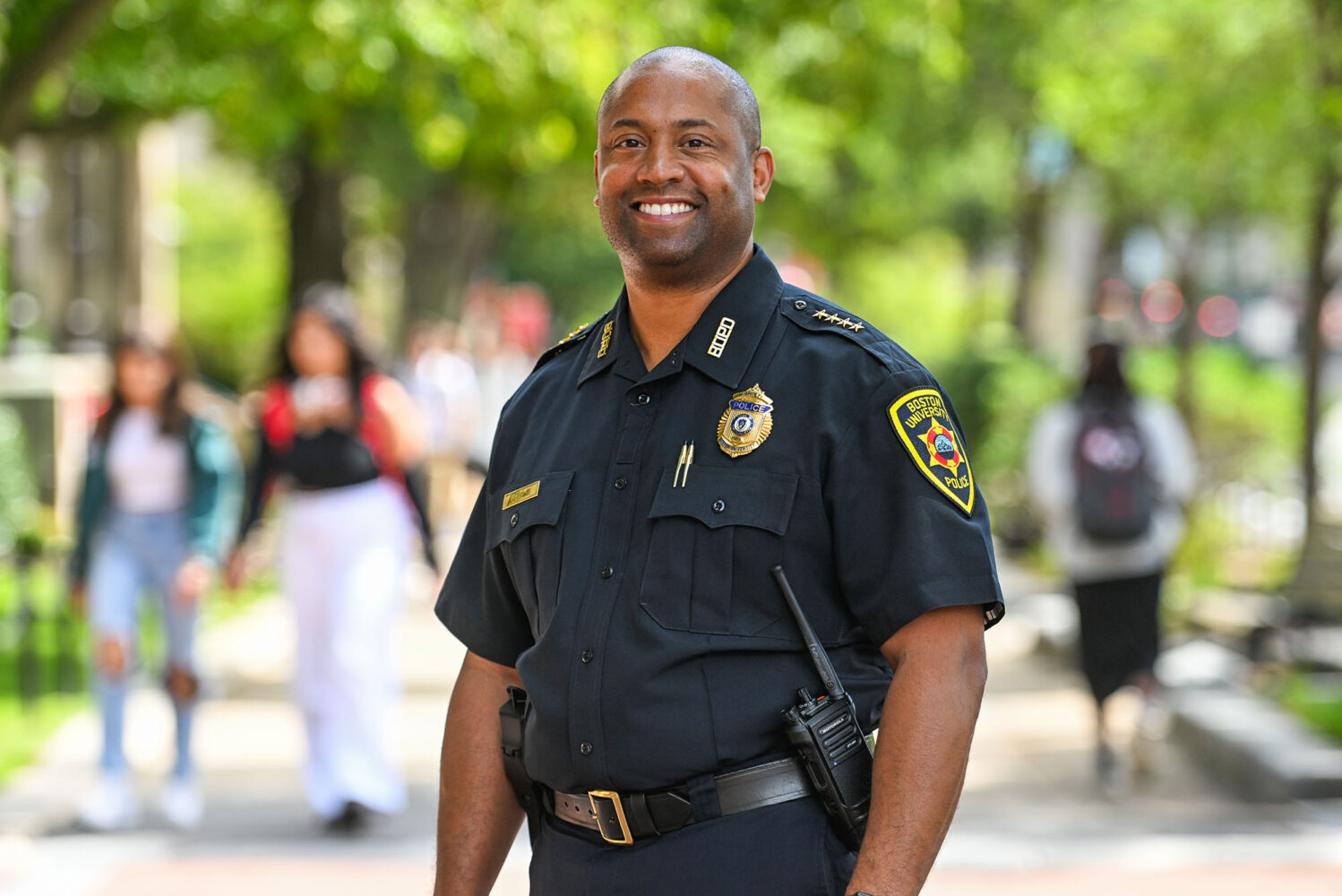 Photo: Robert Lowe, a bald black man wearing a Boston University Police Department uniform, smiles and poses on a busy sidewalk lined with greenery. Blurred pedestrians walk behind him.