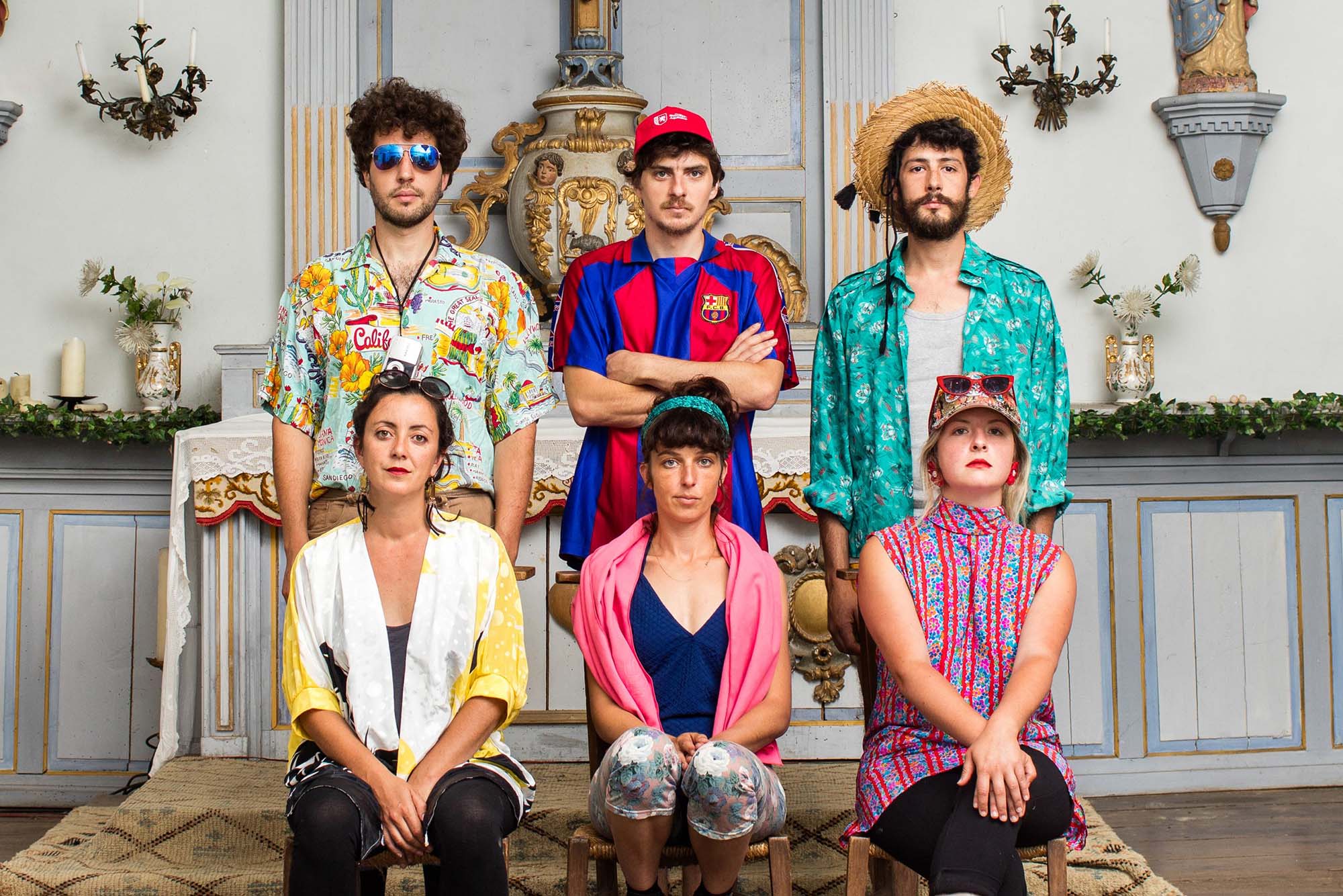 Photo: A group of six people wearing brightly-colored shirts and hats poses for a photo. Three sit while the other three stand behind them. They all wear neutral expressions as the background displays an ornate, European-inspired background.