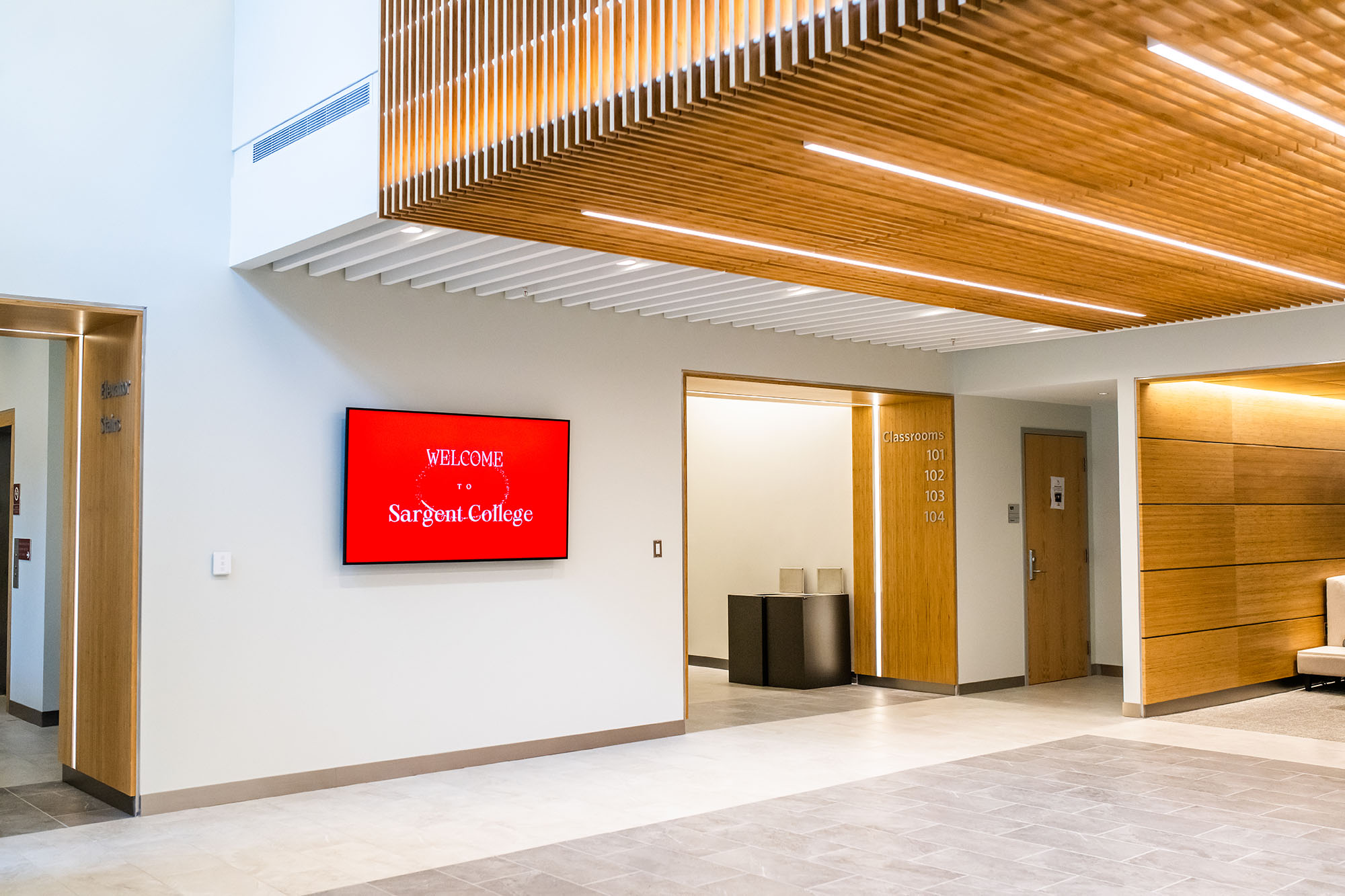 Photo: A bright, modern room with wooden accents is shown. A screen on the wall has a red display that reads in white text "Welcome to Sargent College".