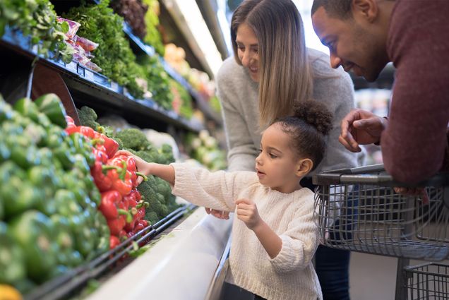 Photo: A pre-school age girl helps her parents pick out veggies in the produce section at the grocery store. She is reaching for a red bell pepper. Her mother is Caucasian and her father is African-American.