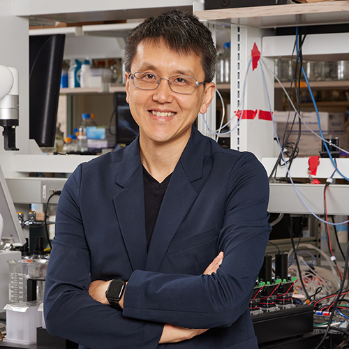 Photo: Wilson Wong, a man wearing glasses, a black shirt, and navy blue blazer, smiles as he poses with arms crossed in a cluttered electrical engineering lab.