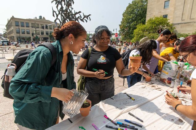 Photo: Two darker-skinned young women looks at and hold up small succulent plants from a long white table. They stand in Marsh Plaza at Boston University.