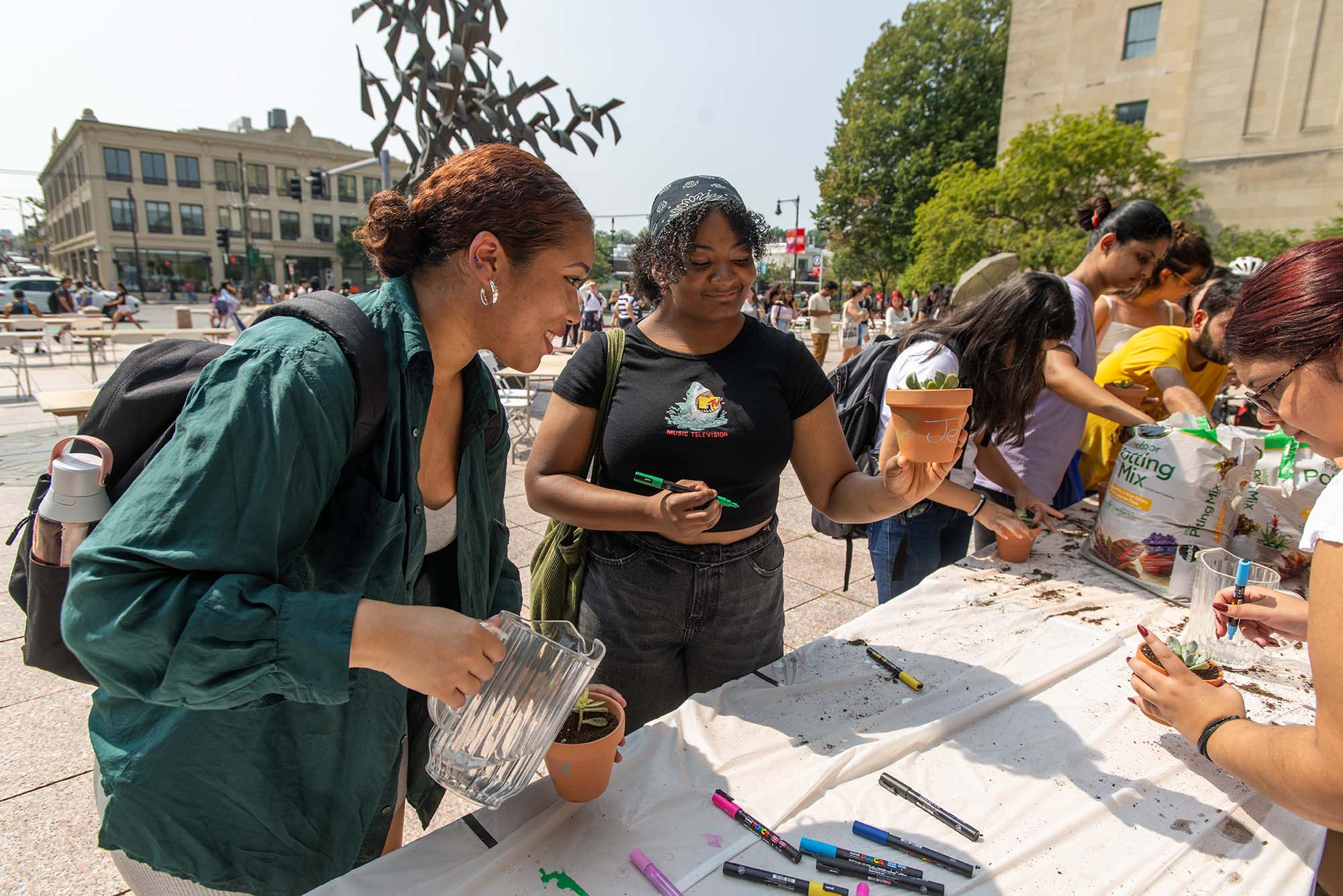 Photo: Two darker-skinned young women looks at and hold up small succulent plants from a long white table. They stand in Marsh Plaza at Boston University.