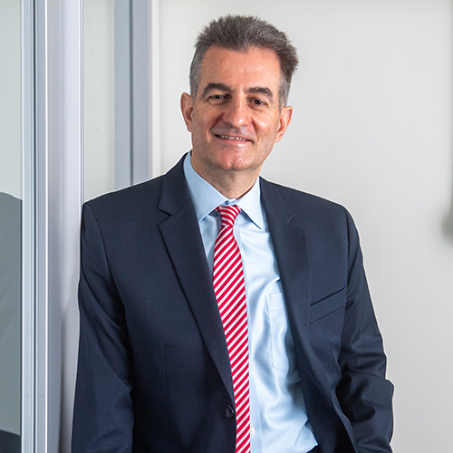 Photo of ENG professor Ioannis Paschalidis, leaning against a glass door and slightly smiling. He wears a dark blue suit, light blue shirt, and red and white striped tie. He is a light-skinned middle-aged man with short brown and gray hair.