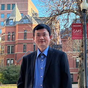 Photo: Yuwei Fan, a Southeast Asian man wearing a blue collared shirt and black jacket, smiles and stands outside a large brick Brownstone building.