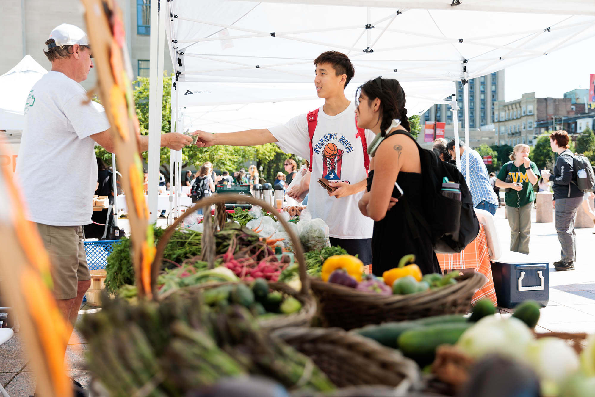 Photo: Two students with dark hair pay a man at a farmers market stand. They are under a white tent, the day is bright, and various vegetables lay in brown wicker baskets.