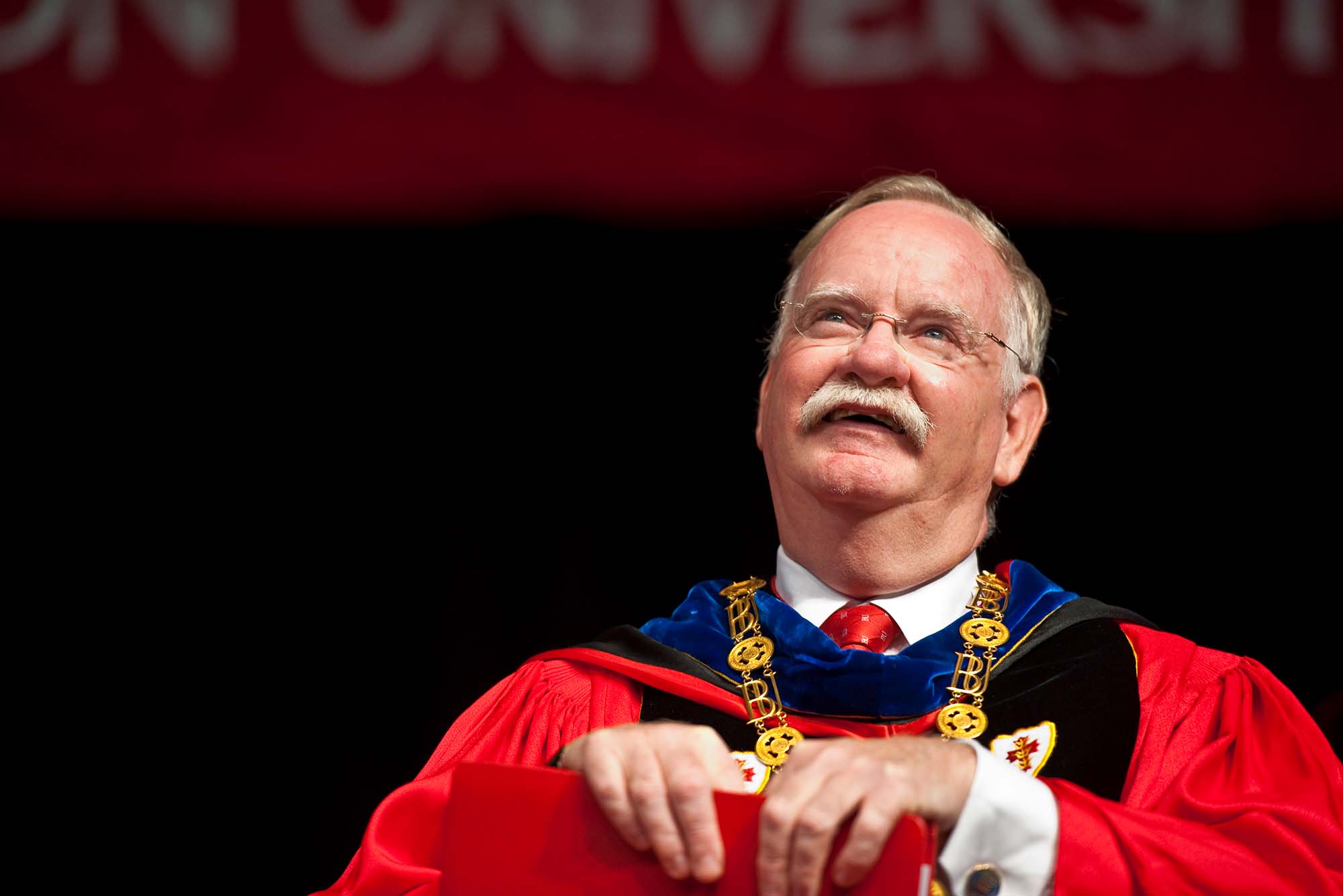 Photo of President Robert Brown in red ceremonial regalia. He has a blue color and wears a gold BU necklace. He is seated, and balances a red folder in his lap and smiles as he looks up into the middle distance. Brown is an older White man with glasses and short gray hair and a mustache.