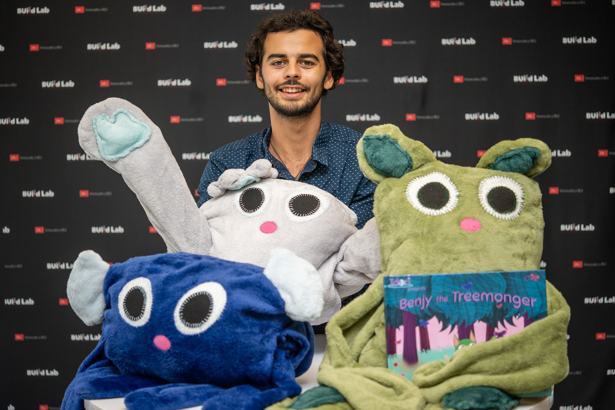 Photo: A man with a button-up shirt sits behind three stuffed creatures, all with huge eyes and long limbs. They are characters from the children book that one of the creatures holds, a book for sustainability for kids.