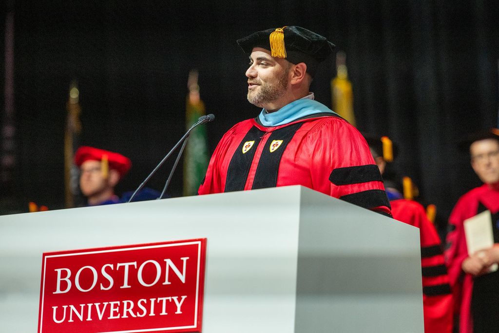 Photo: Jason Campbell-Foster stands a a podium with BOSTON UNIVERSITY plaque wearing graduation ceremonial garb. He is the middle of talking.