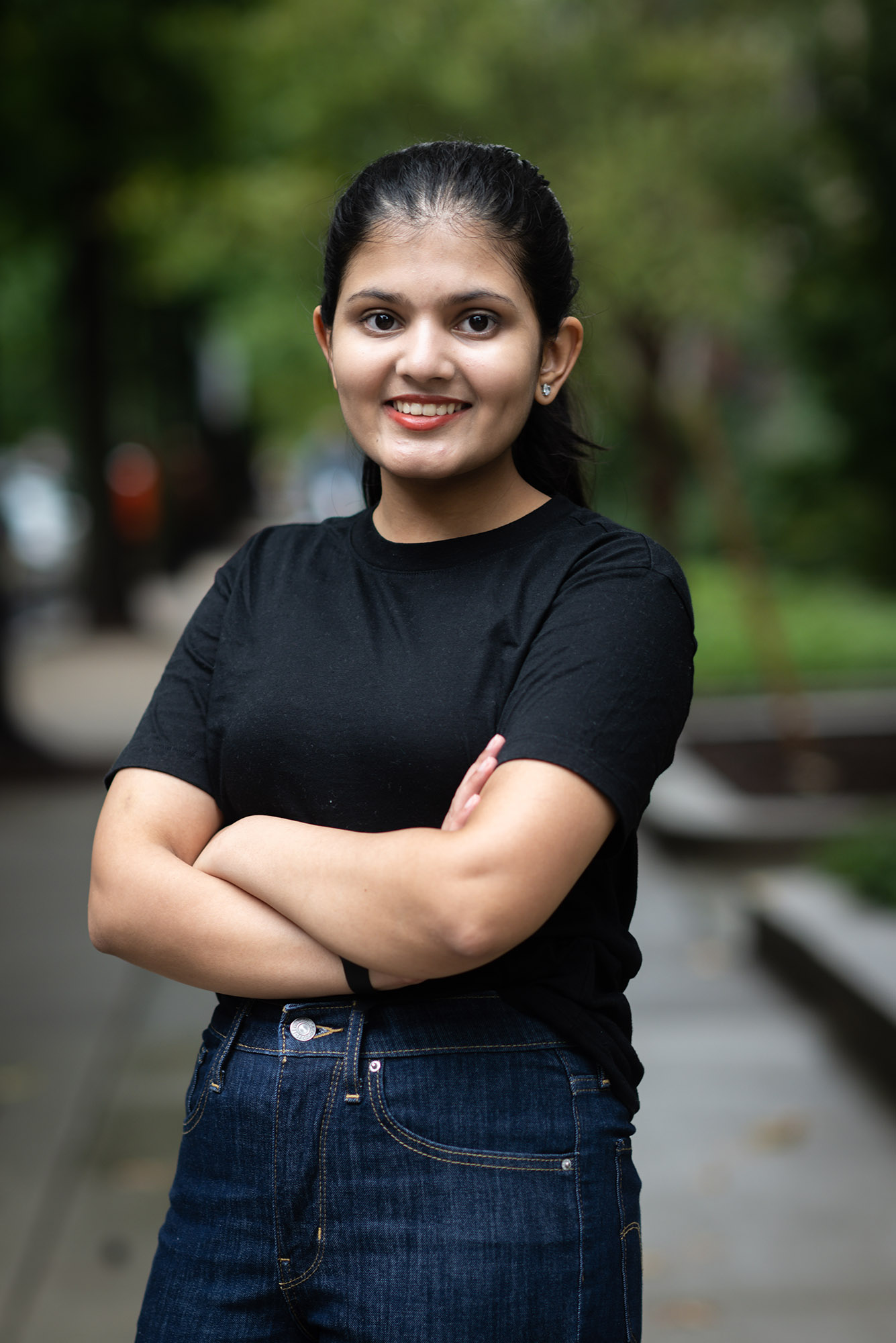 Photo: A young woman with her arms crossed. smiling for the camera. She has her hair pulled back into a ponytail and she wears a black t-shirt and dark jeans. The blurred background behind her is of sidewalk and green foliage.
