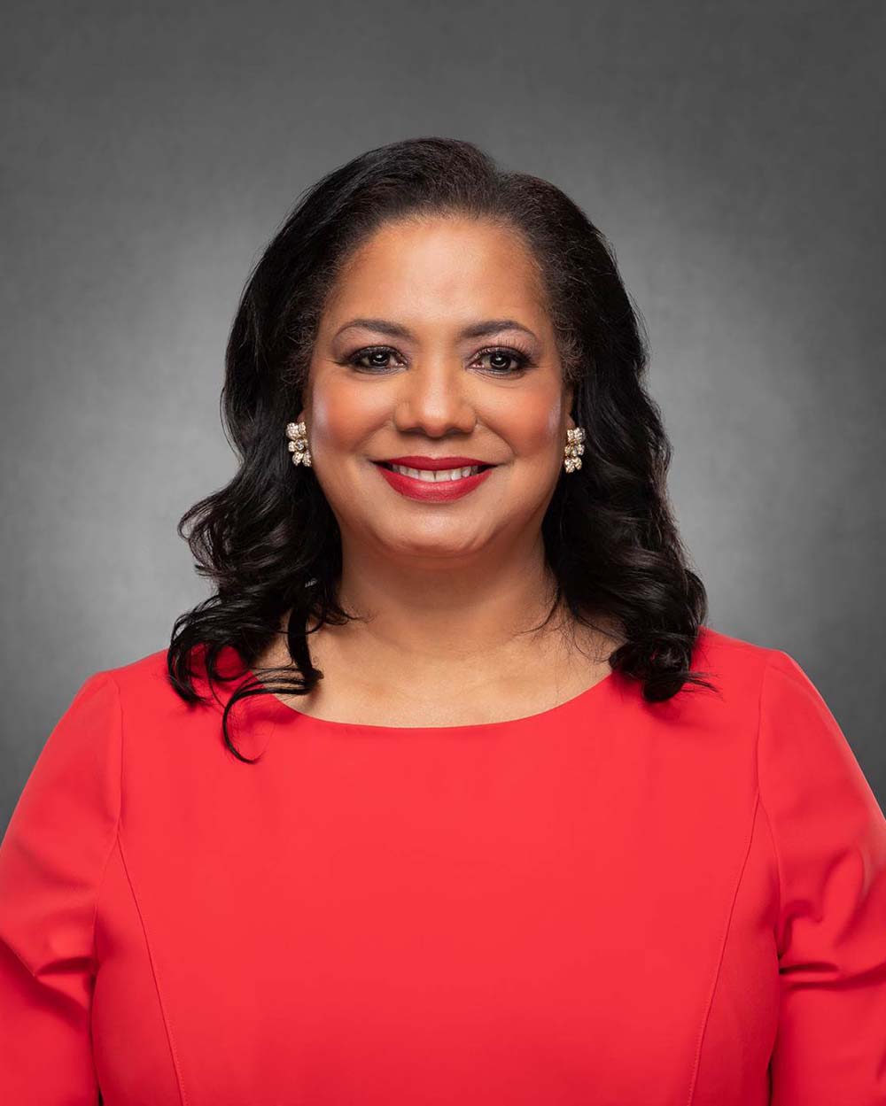 Photo of Antoinette “Tonie” Leatherberry (ENG’85), a middle-aged Black woman with shoulder length, slightly curled dark hair. She smiles, wears read lipstick, and earrings in the shape of a flower. She wears a pinkish-orange dress and stands in front of a dark gray background.