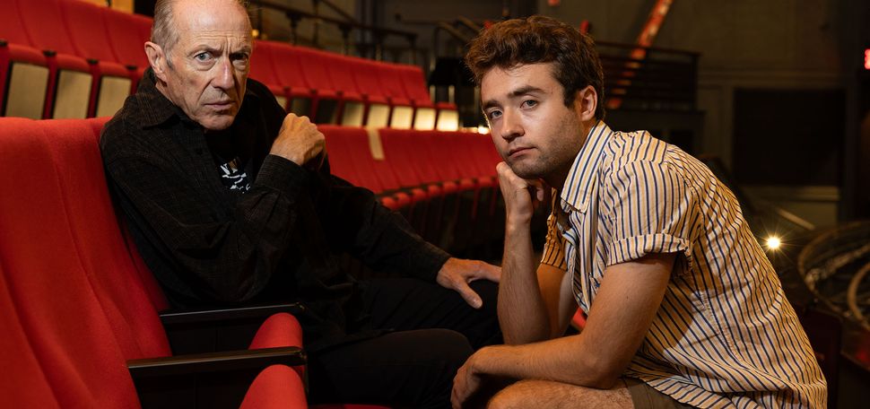 Photo: Will Lyman (CFA’71), an older white man, sits in the audience chairs as he looks at the camera with a side eye, posed strong. Jesse Kodama (CFA’25), a younger white man with short brown hair, sits across from him, holding his head in his hand.