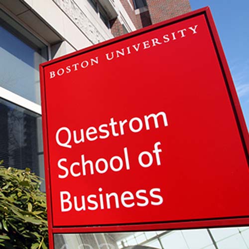 Photo of a red sign with white text that reads "Questrom School of Business." A building and bushes are seen in the background.