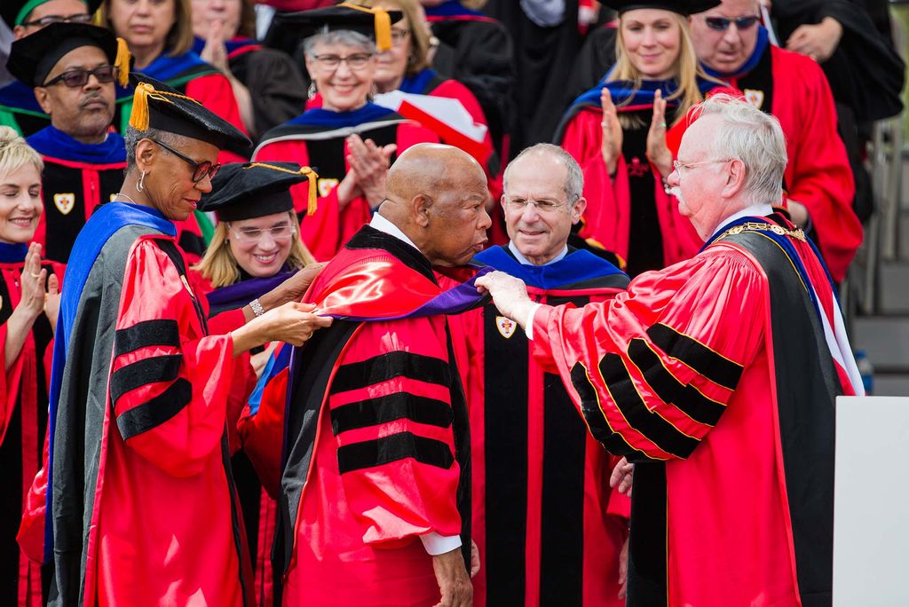 Photo of Boston University President Robert A. Brown, right, and Trustees Andrea Taylor, left, and Kenneth Feld presenting an honorary degree to John Lewis (center) during the 2018 Boston University Commencement on Nickerson Field on May 20, 2018. Each person wears the ceremonial red regalia with blacks tripes on the sleeve. Andrea Taylor is a middle-aged Black woman with glasses who smiles as she helps with Lewis' hood. Civil rights activist John Lewis is an older, bald Black man. Feld and Brown are older White men with glasses. Other faculty in the background look on and clap and smile.