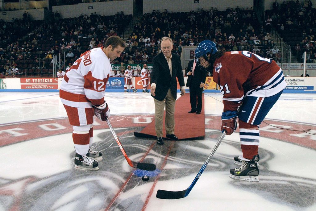 Photo of president brown Boston University President Robert Brown dropping the puck to open the Terrier Men's Ice Hockey 2005-2006 season. He stands on the ice in a suit and leans over to drop the puck. Two players in full hockey gear, minus helmets, stand on the ice at the ready.