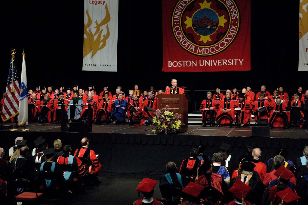 Photo of Robert Brown's Inauguration Ceremony in 2006. Brown stands at the podium in regalia and addresses the crowd. Other faculty and staff are seen seated on stage in regalia and a large Boston University banner hangs behind the stage. People in regalia are seen seated on the ground level.