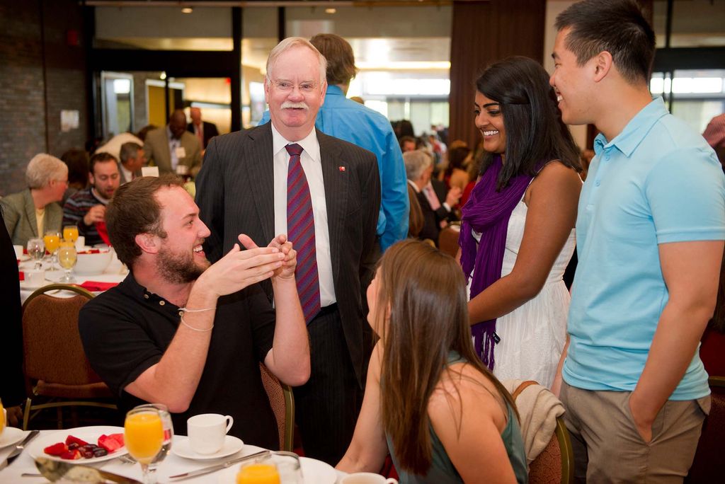 Photo of Class of 2011 Seniors Ray Curran (SAR) sitting at left, Missy Goldberg (CAS), sitting at right, Eddie Chen (SAR) in teal, and Shilpa Reddy (SAR) chat with President Brown before the start of senior breakfast at Metcalf Ballroom May 6, 2011. Curran is a young white man in a black polo who smiles as he gestures with his hands. Goldberg is a white woman with long hair who turns to look at Brown standing behind her. Reddy is a young woman of color in a white dress who smiles. And Chen, in a teal polo, is a young AAPI man. Brown wears a suit and maroon tie.