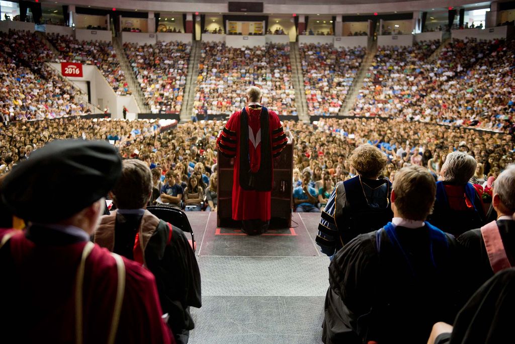 Photo taken from behind of Brown in the red matriculation ceremonial regalia as he speaks to the new class of 2016 during matriculation inside Agganis Arena. The backs of other faculty members in robes and hats are seen from behind in the foreground, while the arena is filled with students.