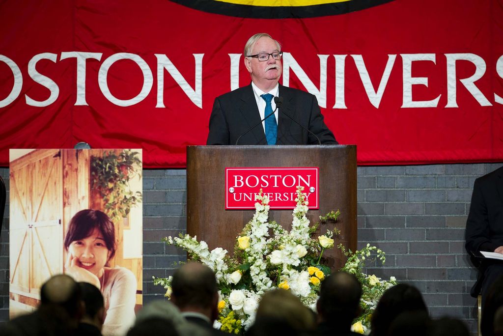 Photo of President Robert Brown standing at a wooden Boston University podium and addressing the audience during the memorial service for Lu Lingzi April 22, 2013 in the GSU Ballroom. Lu, who was from China, was killed by the Boston Marathon bombing. A large photo of Lu is seen at bottom left. Flowers are seen in front of the podium and the back of people's heads in the audience are seen in the foreground.