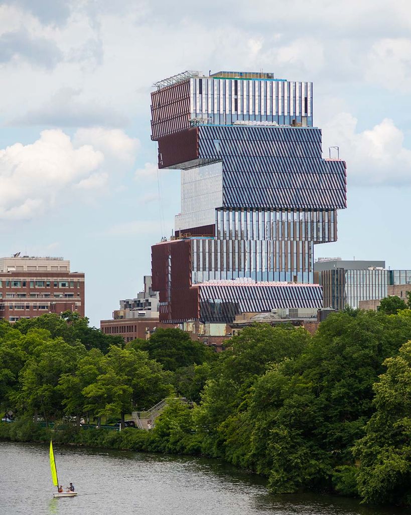 Photo of the Boston skyline as seen from the BU bridge. The reflective blue and reddish Center for Data Science features prominently at right. Bright green summer trees line the Charles River and the sky is a bit overcast. On the river, a small sunfish sailboat with a yellow-green sail is seen. 