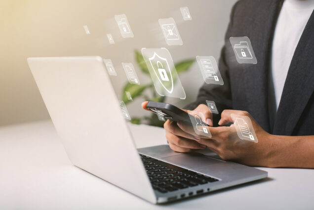 Photo: A person holds their phone while on their laptop in a stock photo. Icons of locks and cyber images are overlayed on the photo.