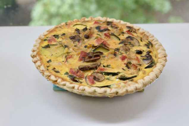 Photo: A bright image of a crusted quiche, with various amounts of vegetables like mushrooms, tomatoes, eggplant, and more. The round quiche sits on a bright, white table.