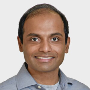 Photo: Headshot photo of a tan man wearing a grey collared shirt. He smiles and poses in front of white background.